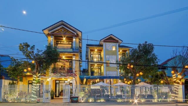 Three-story Hoi An building with balconies, lit lanterns, and trees, behind a white picket fence at dusk.