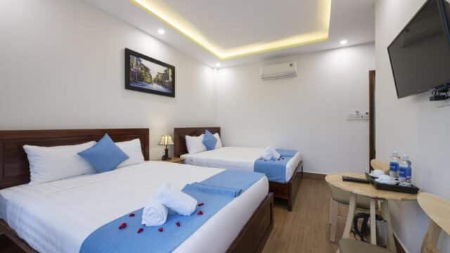 Modern Hoi An hotel room with two beds, blue accents, wall art, TV, and a small table with water bottles.