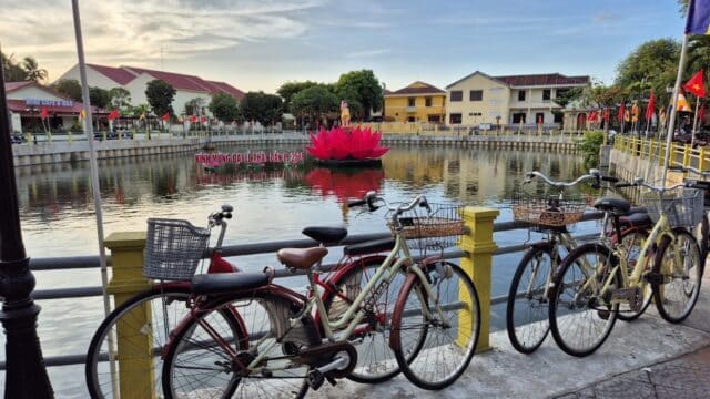 Several bicycles are parked by a yellow railing near Hoi An’s calm lakeside, with buildings and a lotus statue in the background.