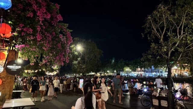 A lively Hoi An riverside street at night with people walking, lanterns, flowers, and colorful lights reflected on the water.