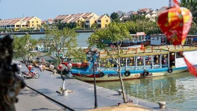 Colorful boats docked along a Hoi An riverside walkway with trees, buildings, and a red lantern in the foreground.