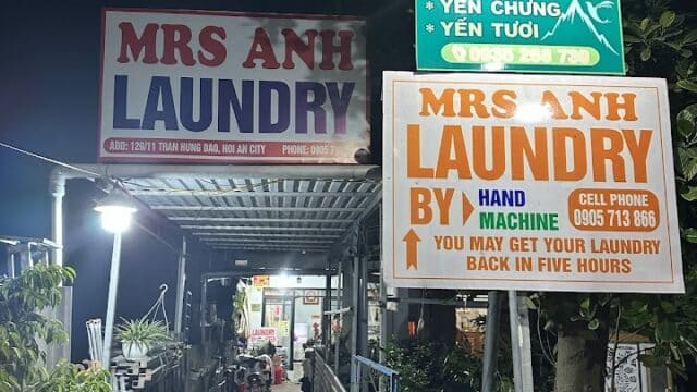 Outdoor night scene in Hoi An with two Mrs Anh Laundry signs, a street sign, plants, and items by a covered walkway.
