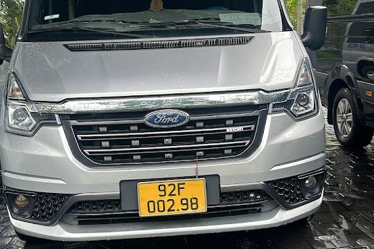 A silver Ford van with a yellow license plate is parked on wet Hoi An pavement under a roof with trees in the background.