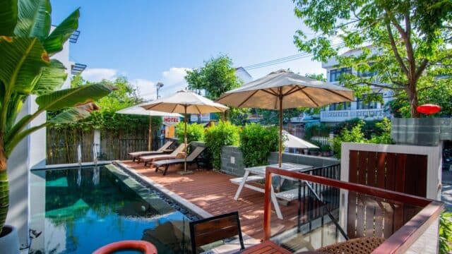 Sunny poolside area with lounge chairs, umbrellas, and lush greenery at a small Hoi An hotel under a clear blue sky.