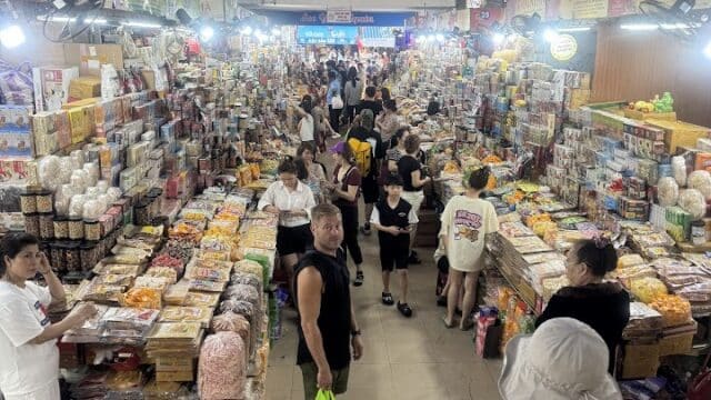 Bustling indoor Hoi An market with people shopping at stalls filled with packaged goods and snacks, seen from above.