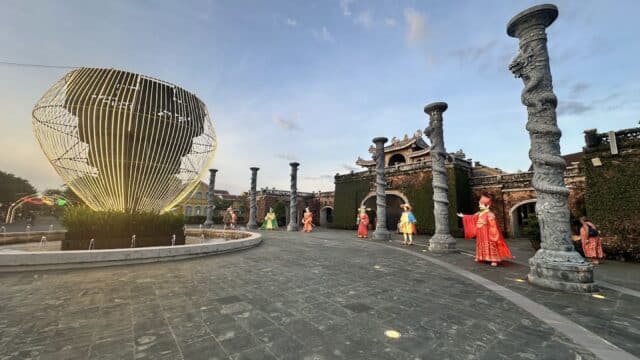 People in traditional costumes stand near tall columns and a modern sculpture in an open Hoi An courtyard at sunset.