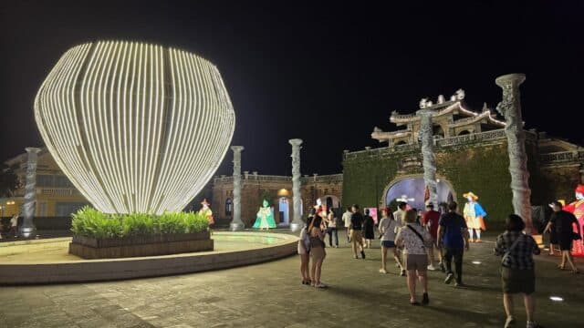 People walk at night near a large, glowing light sculpture and an ornate Hoi An building with columns and arches.
