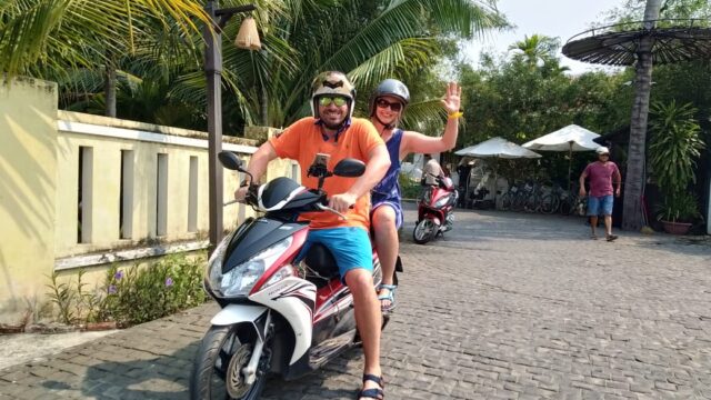 Two people wearing helmets ride a scooter on a sunny Hoi An street, smiling; the passenger waves. Palm trees and people are in the background.
