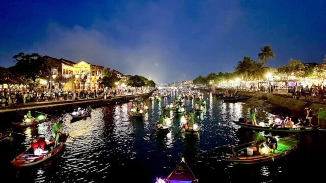 Colorful lantern-lit boats float on Hoi An’s river at night, with crowds and lights glowing along the riverbank.