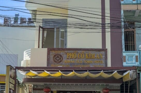 Three-story yellow Hoi An hotel with a brown Pho Co Kim An sign and flower stands by the entrance, under a striped awning.