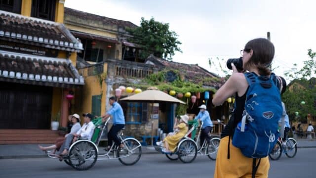 A woman with a camera and blue backpack photographs people riding cyclos on a Hoi An street lined with old buildings.