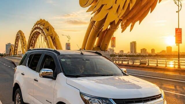 A white SUV parked on a bridge in Hoi An with a large yellow dragon sculpture in the background at sunset.