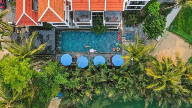 Aerial view of a pool by a Hoi An building with red roofs, blue umbrellas, palm trees, and a river nearby.