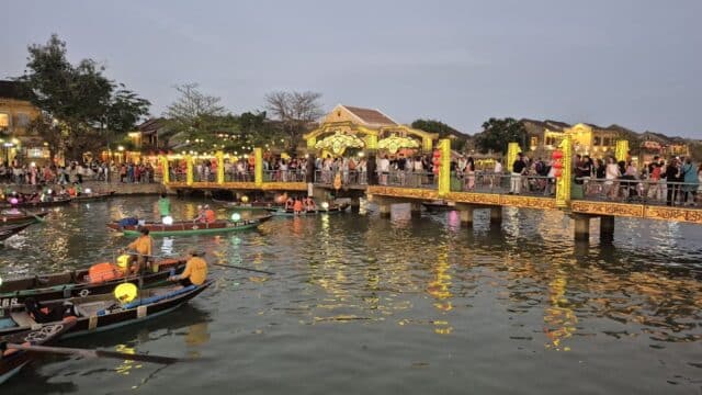 Crowds on a decorated bridge over Hoi An’s river, with boats and lanterns glowing in the evening.