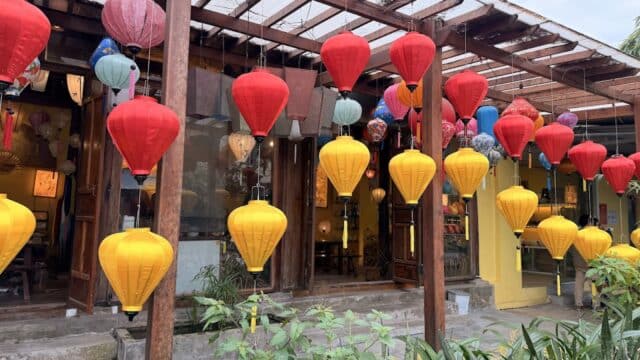 Colorful red and yellow lanterns in Hoi An hang outside a building with glass windows and wooden beams, surrounded by plants.