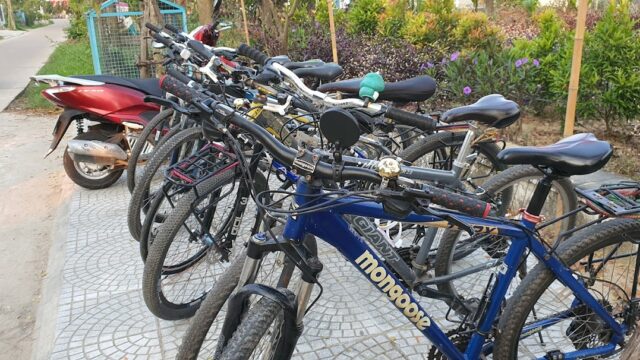 Several bicycles and a red scooter are parked on a sidewalk in Hoi An, near a street lined with greenery and purple flowers.