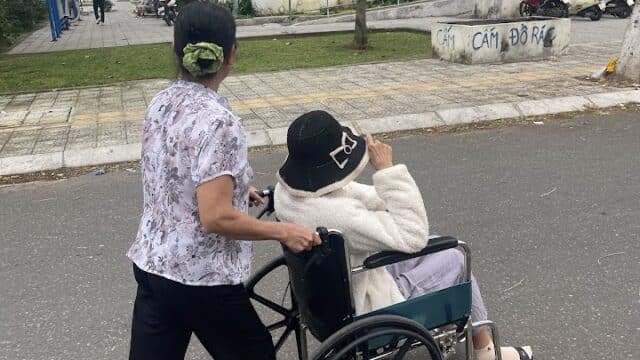 A woman pushes another woman in a wheelchair along a street near an apartment building in Hoi An on a cloudy day.