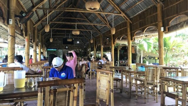 Open-air bamboo restaurant in Hoi An with wooden tables, lively conversations, pitchers of drinks, and lush greenery outside.