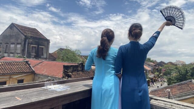 Two women in blue dresses stand on a Hoi An balcony, one holding a fan, overlooking rooftops and a street under a partly cloudy sky.