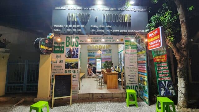 A brightly lit Hoi An bus and limousine ticket office at night with green stools outside and signs displaying services and schedules.