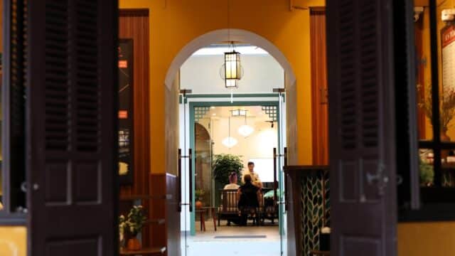 Two people sit at a table in a bright Hoi An room, seen through an arched doorway and open double doors.