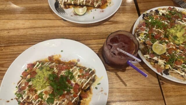 Three plates of loaded nachos with toppings, plus a reddish drink with ice and two straws on a wooden table in vibrant Hoi An.
