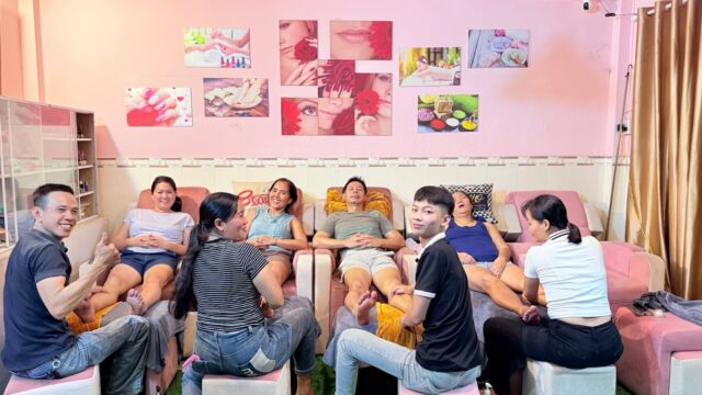 Eight people relax in a brightly lit Hoi An salon, chatting and smiling while getting pedicures on stylish pink chairs.