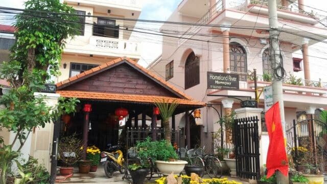 Two Hoi An houses with balconies, a small garden, bicycles, and a red Vietnamese flag line a street beneath power lines.