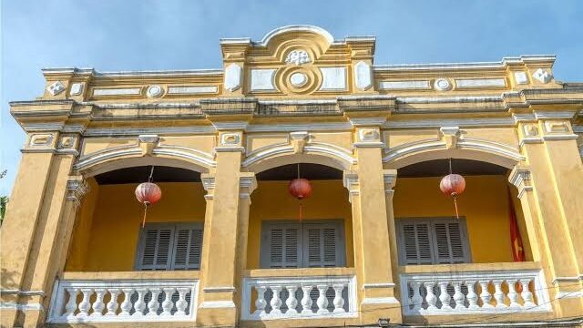 Yellow colonial-style building in Hoi An with arched windows, balconies, and red lanterns hanging under the arches.