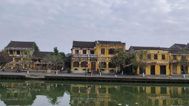 Old yellow buildings line a calm riverside street in Hoi An, reflected clearly in the water under a cloudy sky.