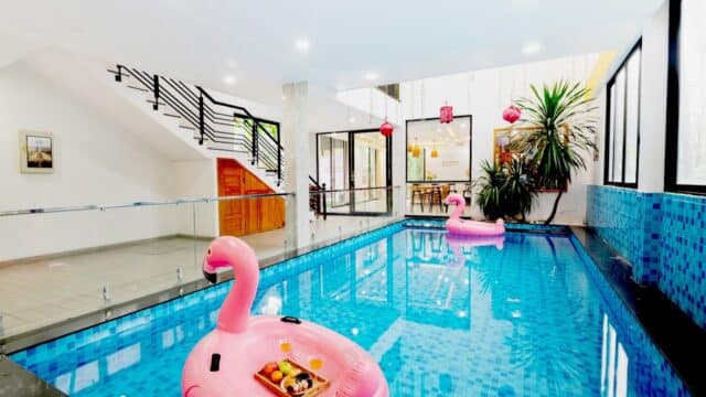 Indoor pool with blue tiles, two pink flamingo floats, plants, large windows, and a modern staircase evoke a Hoi An-inspired retreat.