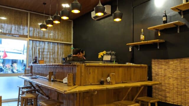 A cozy bamboo-themed bar in Hoi An, with stools, warm lighting, and bottles on shelves. Two people stand behind the counter.