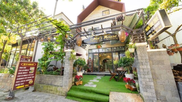 Entrance to Lynas House restaurant in Hoi An features green turf, lush plants, flowers, and welcoming signs in a bright outdoor setting.