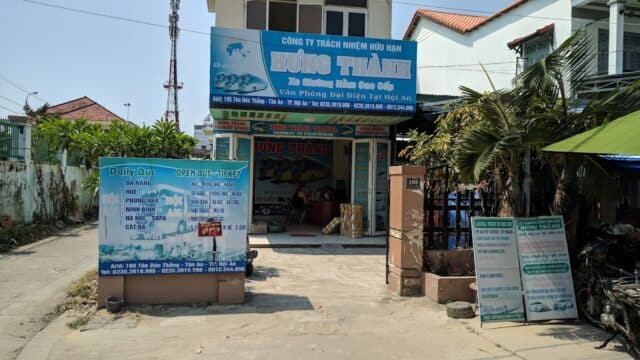 Bus ticket office in Hoi An, Vietnam with blue signage, ticket prices displayed, and a few people inside near a desk.