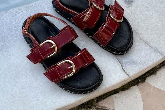 A pair of red buckle sandals, inspired by Hoi An, placed on a marble surface beside a swimming pool.