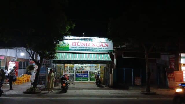 A pharmacy storefront in Hoi An lit up at night, with people, scooters, and trees lining the lively sidewalk.