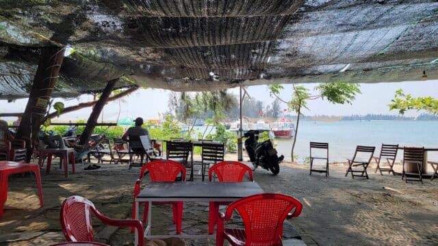 Shaded outdoor café in Hoi An with red chairs facing the river, boats, and trees in the background under a net canopy.