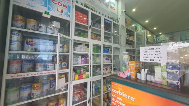 Pharmacy interior in Hoi An with shelves of medicine, formula cans, and a sign reminding customers to keep a 2-meter distance.