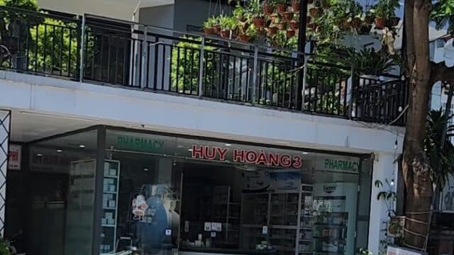 A glass-fronted pharmacy named Huy Hoang 3 in Hoi An, with plants on the balcony above and a tree on the sidewalk in front.