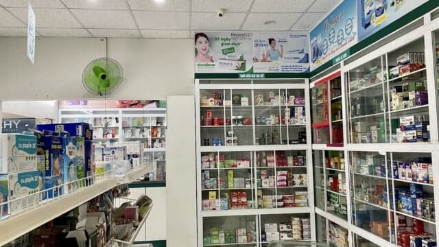 Shelves filled with medicines and products inside a brightly lit Hoi An pharmacy, featuring glass cabinets and advertising posters.