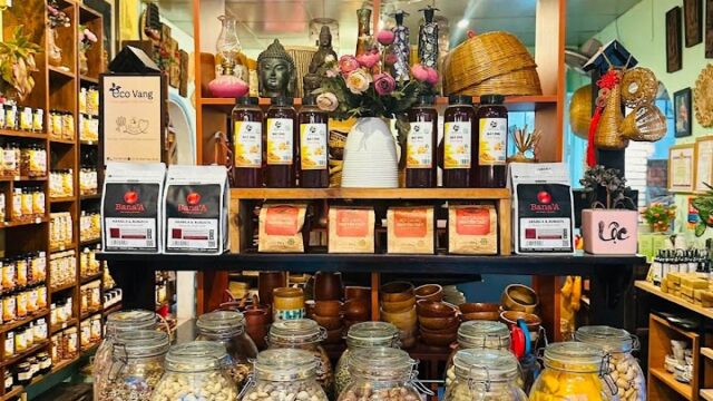 Six large glass jars filled with beans and nuts sit on a black table in a cozy, well-lit Hoi An shop lined with shelves.