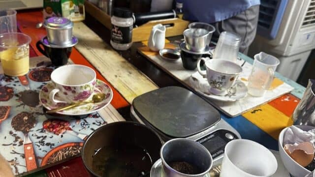 A person prepares coffee at a colorful table in Hoi An, surrounded by cups, filters, and various brewing tools.