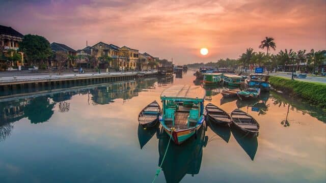 Colorful boats on a calm river at sunset in Hoi An, with historic buildings and palm trees lining the riverbank.