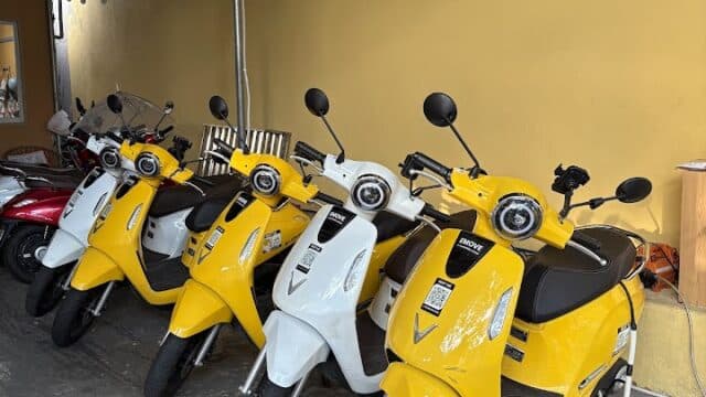 Four yellow and white scooters are parked in a row by a yellow wall under string lights in charming Hoi An.