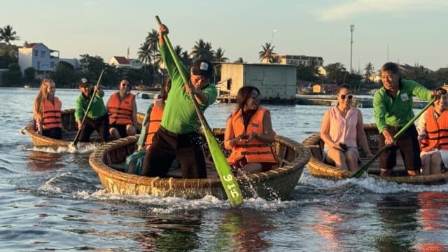 People wearing life jackets paddle round basket boats on a calm river in Hoi An, with palm trees and buildings in the background.