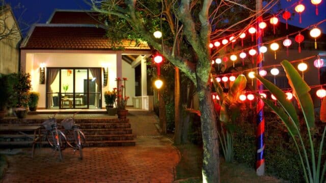 A cozy Hoi An house at night with red lanterns, bikes outside, trees, and warm inviting lights illuminating the path.