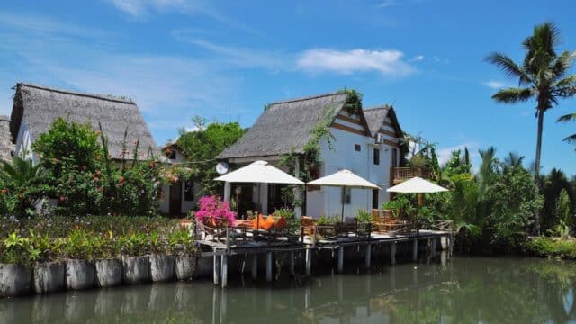 Thatched-roof houses with outdoor seating and umbrellas beside a calm river in Hoi An, surrounded by lush greenery and flowers.