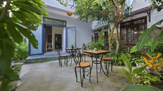 Outdoor patio with round tables, chairs, and lush green plants in a garden setting next to a building in charming Hoi An.