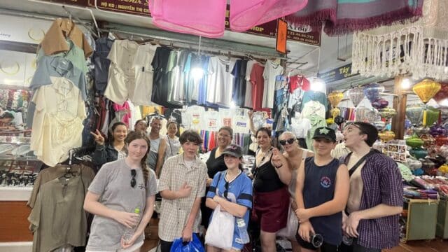 A group of people pose and smile inside a busy Hoi An clothing market filled with hanging shirts and souvenirs.