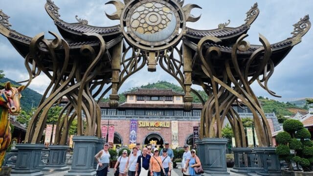 A group of people poses in front of Sun World’s ornate entrance gate in Hoi An, with a mountain visible in the background.